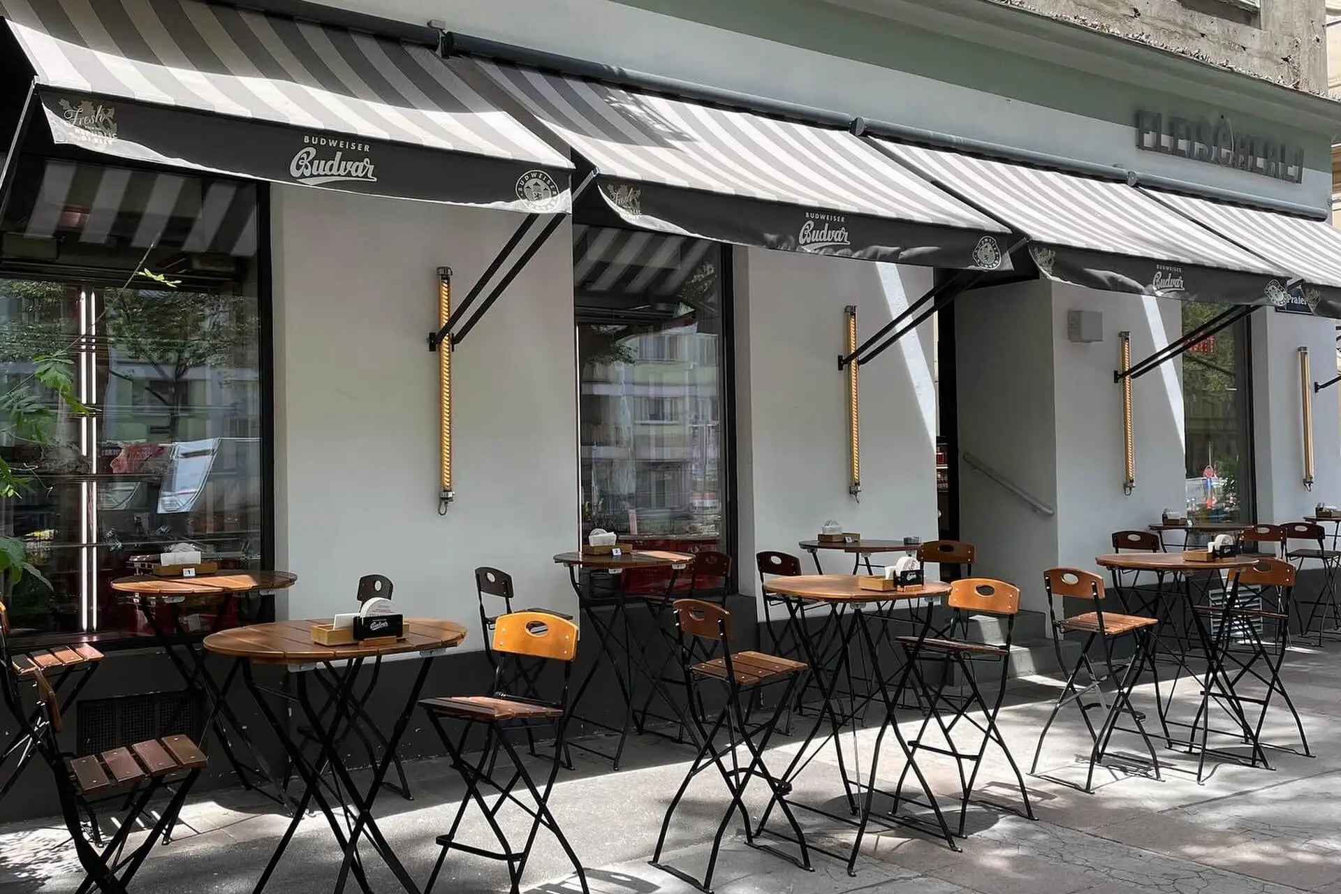Außensitzbereich eines Cafés mit schwarz-weiß gestreiften Markisen, Holztischen und -stühlen und kleinen Pflanzen neben den Tischen an einem sonnigen Tag. Das Café ist leer, und das Sonnenlicht wirft Schatten auf den Bürgersteig.