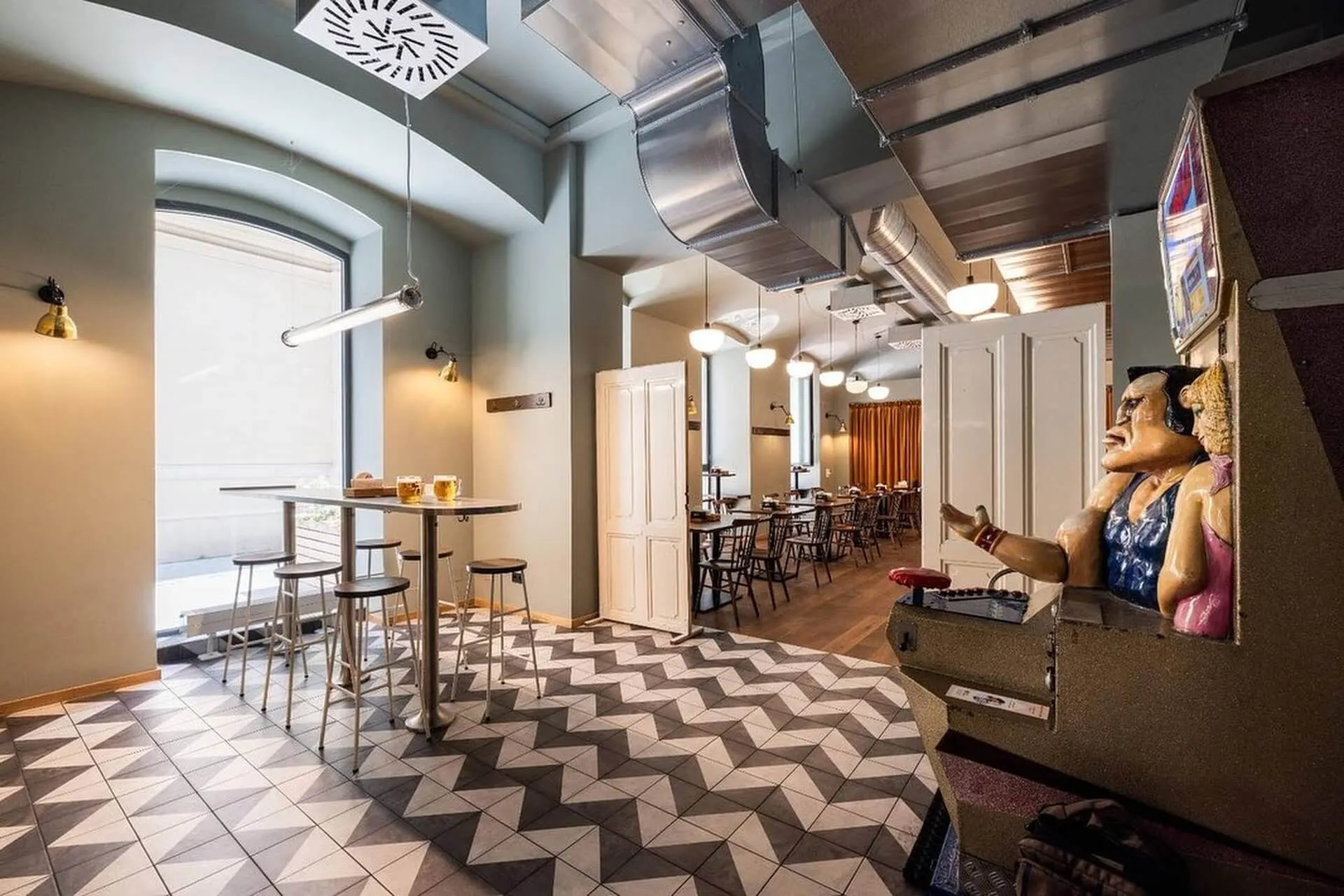 Ein modernes, gut beleuchtetes Café mit geometrischen Bodenfliesen, hohen Tischen und Hockern an großen Fenstern, Esstischen im Hintergrund und einem alten Boxautomaten mit einer Boxerstatue auf der rechten Seite.