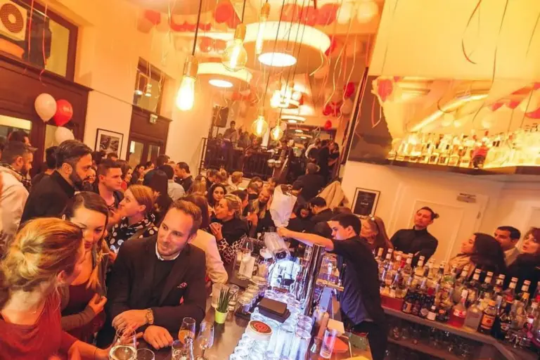 Eine überfüllte, lebhafte Bar mit Menschen, die sich unterhalten und trinken; Barkeeper bedienen am Tresen, und von der Decke hängen festliche rote und weiße Luftballons und Luftschlangen, die eine feierliche Atmosphäre schaffen.
