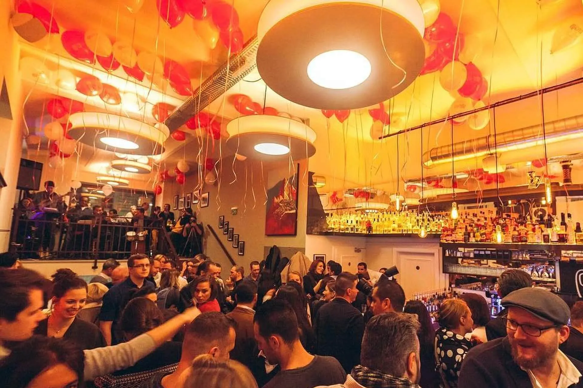 Eine überfüllte Bar mit Menschen, die sich unter warmen Lichtern unterhalten, mit roten Luftballons an der Decke und einer gut gefüllten Bar auf der rechten Seite. Die Atmosphäre ist lebendig und festlich.