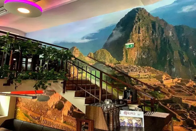 Ein Restaurant mit einer Treppe, Zimmerpflanzen und einem großen Wandgemälde von Machu Picchu, das den Eindruck erweckt, die Treppe führe in die historische Stätte.
