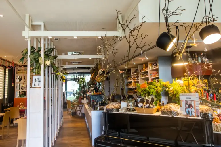 Innenansicht eines gemütlichen Cafés mit einem weißen Trennwand aus Holz, üppig blühenden Pflanzen und einer Theke, auf der Blumenarrangements und Gebäck ausgestellt sind.