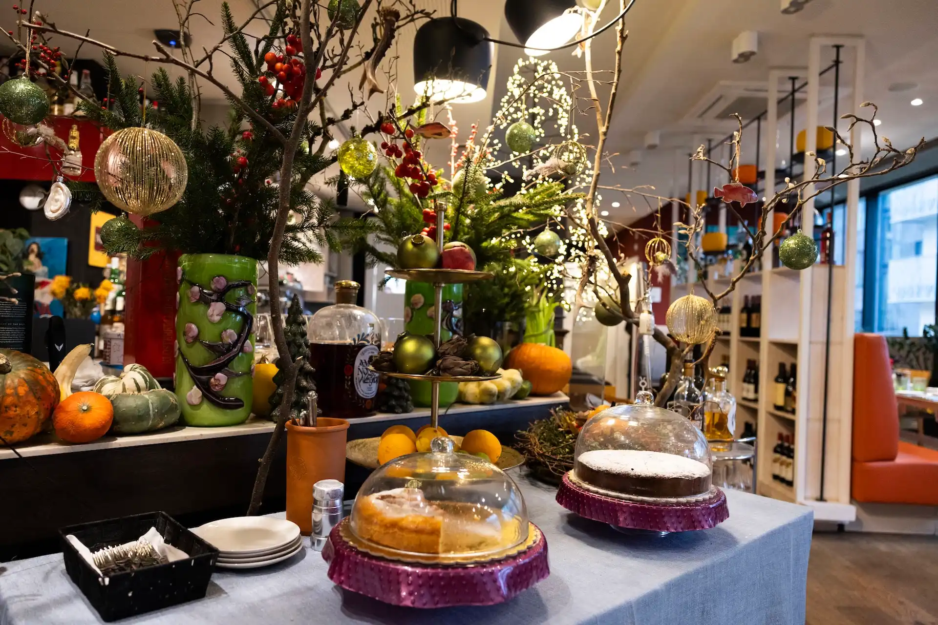 Zwei Tortenständer mit Glashauben auf einem Tisch, geschmückt mit weihnachtlichen Zweigen, Christbaumkugeln und kleinen Kürbissen.