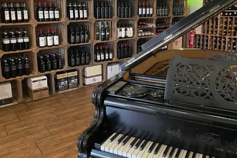 Ein schwarzer Flügel mit verschnörkelten Details steht in einem Geschäft mit Holzfußboden vor Regalen mit verschiedenen Weinflaschen und Kisten.