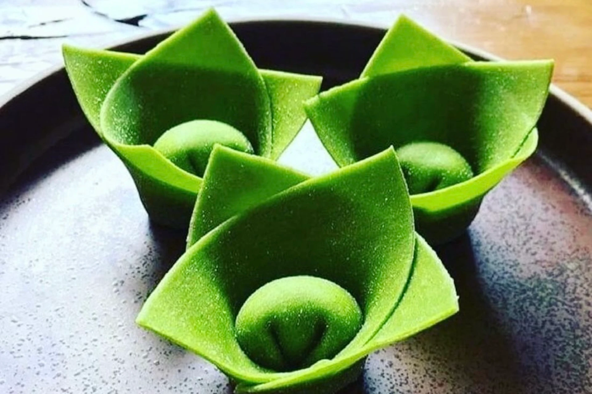 Drei grüne, lotusförmige Desserts sind ordentlich auf einem braunen Teller angeordnet. Die Desserts haben glatte, gefaltete Blütenblätter und eine runde Mitte, die an Lotusblumen erinnern.
