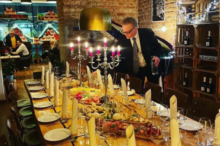 Ein Mann im Anzug schenkt Wein an einem langen, festlich gedeckten Tisch mit Kerzen, gefalteten Servietten und verschiedenem Geschirr in einem gemütlichen, warm beleuchteten und mit Lichterketten und Weinflaschen dekorierten Restaurant ein.