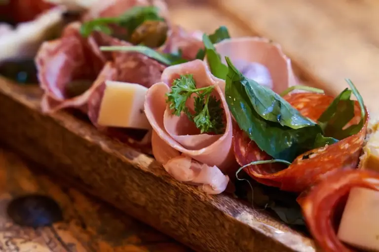 Nahaufnahme eines Holzbretts mit verschiedenen Wurstwaren, darunter gerollte Wurstwaren, Käsewürfel, frisches Grün und mit Kräutern garniert.