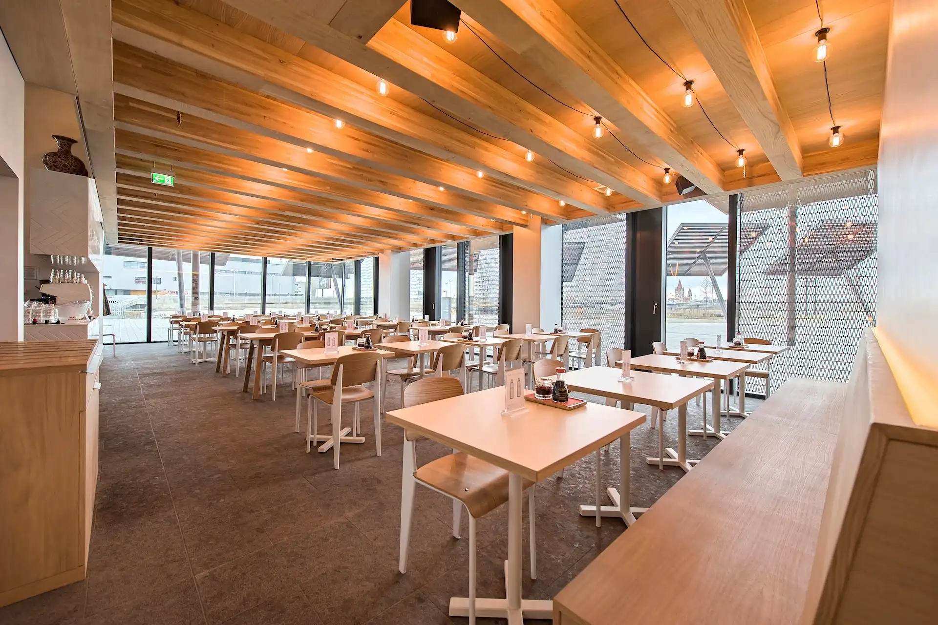 Ein modernes, geräumiges Restaurant mit Holzbalken an der Decke, Hängelampen und großen Fenstern. Helle Holztische und -stühle sind ordentlich angeordnet und schaffen eine klare und einladende Atmosphäre.