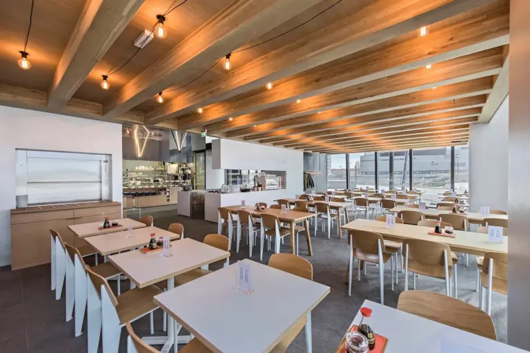 Ra'mien GO DC Tower Moderne Cafeteria mit Holzbalken an der Decke, großen Fenstern und ordentlich angeordneten Tischen und Stühlen. Auf einigen Tischen stehen Flaschen mit Gewürzen. Der Raum ist durch Hängeleuchten gut ausgeleuchtet und verfügt über einen einsehbaren offenen Küchenbereich.