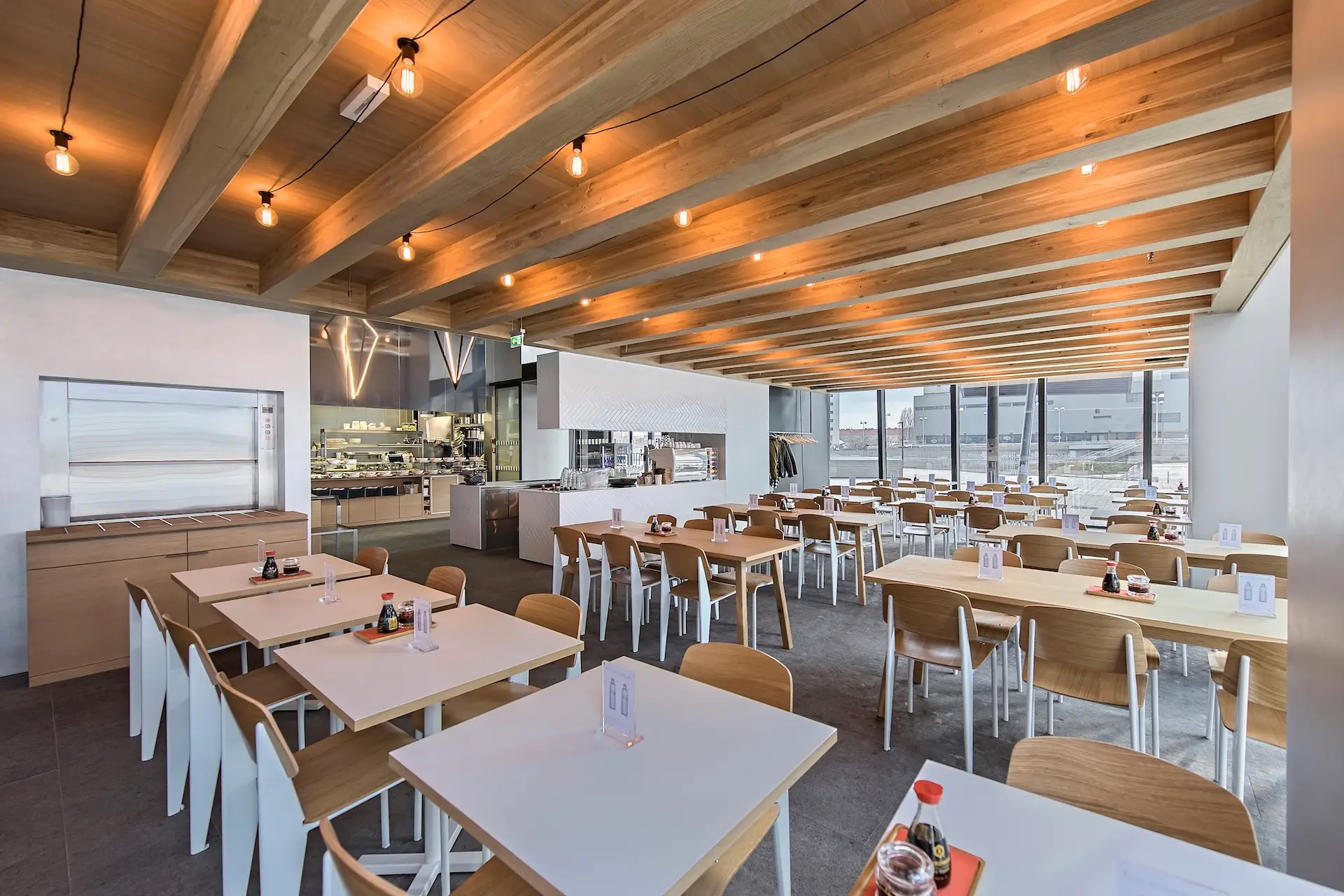 Ra'mien GO DC Tower Moderne Cafeteria mit Holzbalken an der Decke, großen Fenstern und ordentlich angeordneten Tischen und Stühlen. Auf einigen Tischen stehen Flaschen mit Gewürzen. Der Raum ist durch Hängeleuchten gut ausgeleuchtet und verfügt über einen einsehbaren offenen Küchenbereich.