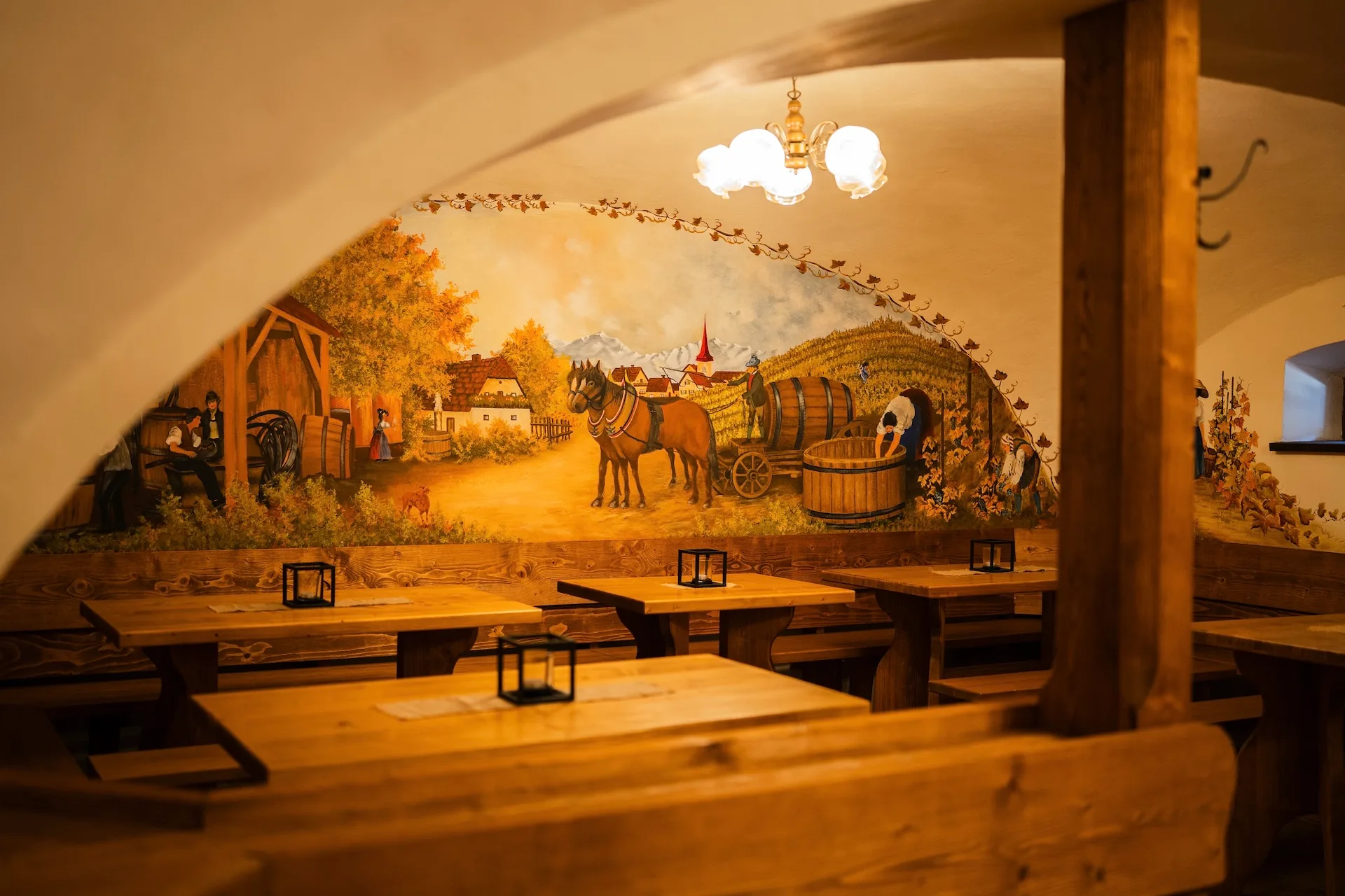 Ein gemütliches, rustikales Restaurantinterieur mit Holztischen und -bänken. Ein Wandgemälde an der gewölbten Wand zeigt eine ländliche Szene mit Pferden, Fässern und Menschen, die unter strahlendem Himmel auf einem Feld arbeiten.