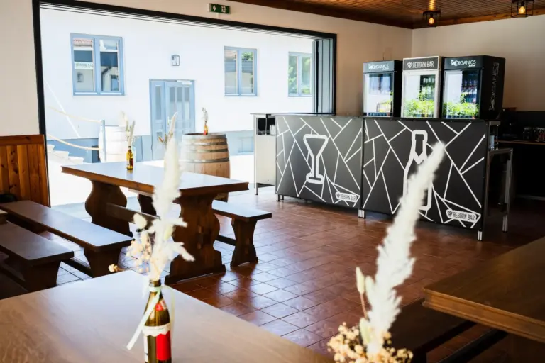 Reborn Bar Moderner Barbereich im Innenbereich mit Holzbänken und -tischen, Trockenblumen in Flaschen, einer dekorativen schwarzen Bartheke und Kühlschränken im Hintergrund. Sonnenlicht strömt durch große Fenster und eine offene Tür herein.