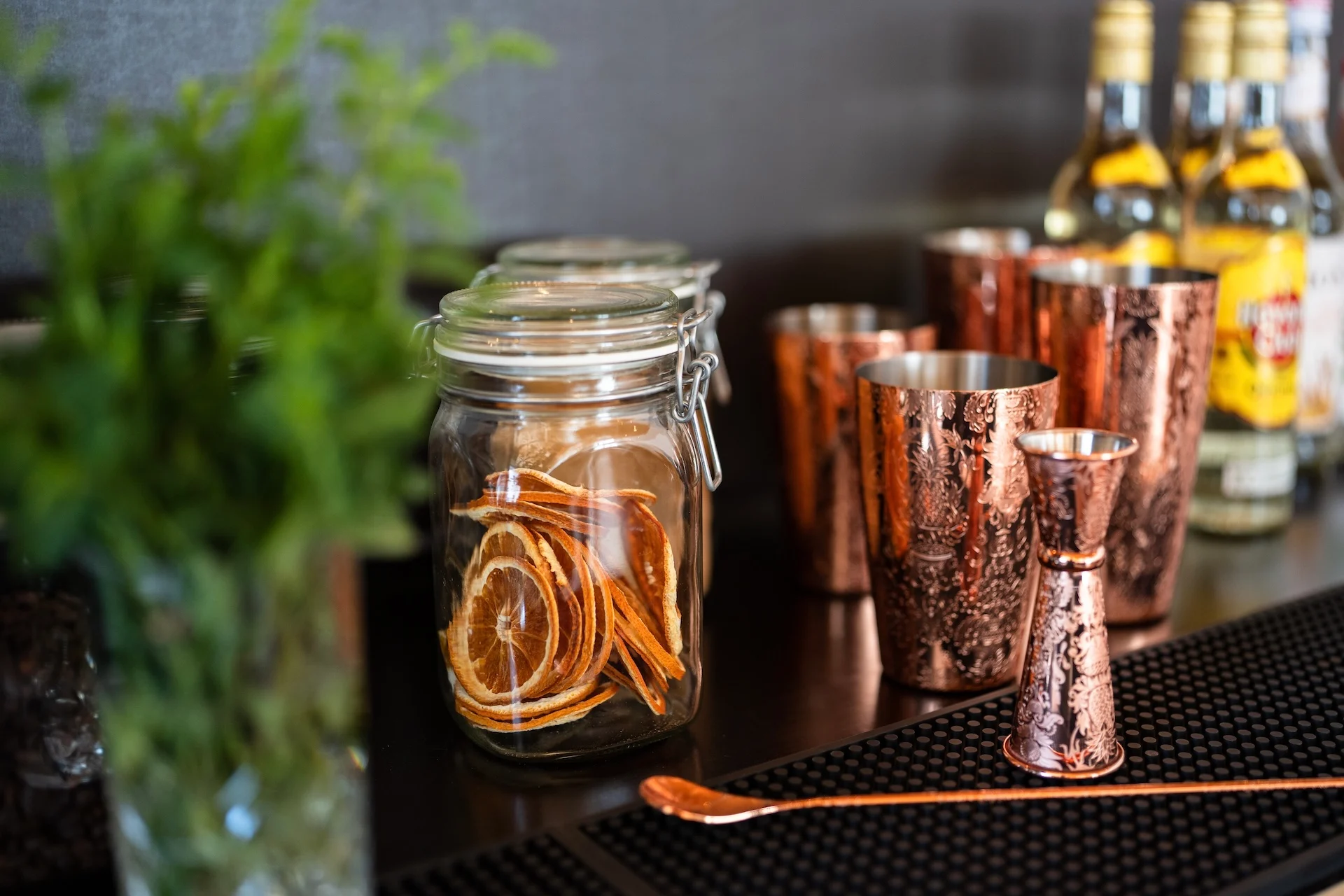 Auf einer Bartheke steht ein Glas mit getrockneten Zitrusscheiben, daneben Kupferbecher, ein Jigger, ein Rührlöffel, grüne Kräuter in einem Glas und im Hintergrund Flaschen mit Spirituosen.