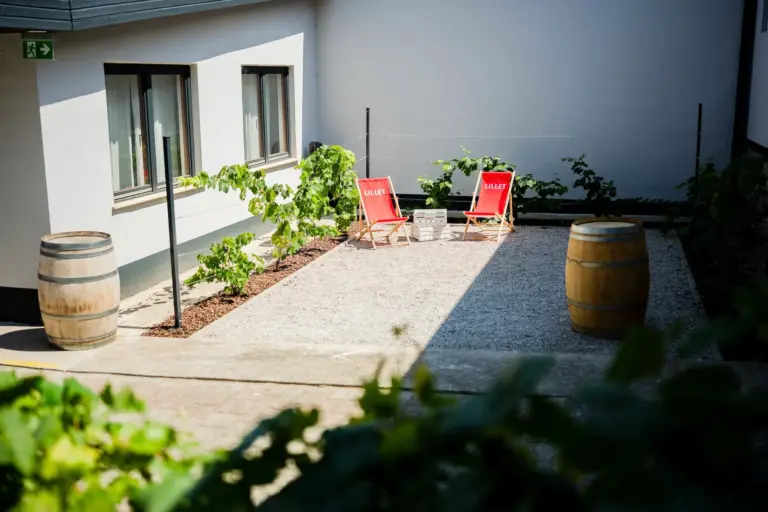 Zwei rote Liegestühle und ein kleiner Tisch stehen auf einer mit Kies bedeckten Terrasse zwischen zwei großen Holzfässern, direkt neben einem weißen Gebäude mit Fenstern und grünen Pflanzen am Rand.