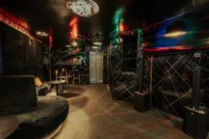 Eine schummrig beleuchtete Lounge mit schwarzen Plüschsitzen, verspiegelten Wänden, bunten Lichtreflexen und kleinen Tischen, die eine stilvolle und moderne Nachtclub-Atmosphäre schaffen.