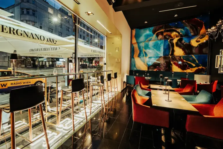 Remember Modernes Café-Interieur mit hohen Hockern an großen Fenstern, farbenfrohen Stühlen an den Tischen und einem lebhaften abstrakten Wandgemälde. Draußen sind die Straße und die Schaufenster der Stadt durch das Glas zu sehen. Warme, einladende Beleuchtung erhellt den Raum.