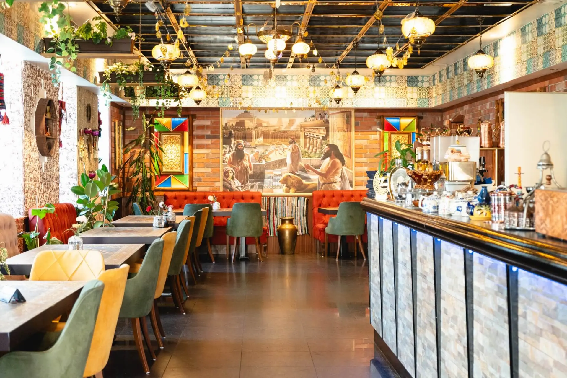 Ein hell erleuchtetes Restaurant mit grünen und gelben Stühlen, einer langen gepolsterten roten Bank, bunten Wandkacheln, Hängelampen, Pflanzen und einer mit Mosaikfliesen verzierten Theke, die eine gemütliche und lebendige Atmosphäre schaffen.
