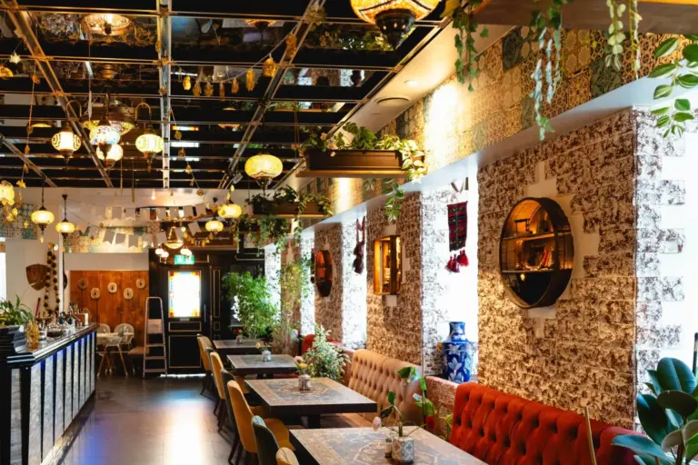 Ein lebhaftes Restaurantinterieur mit gemusterten Wänden, hängenden Pflanzen, runden Spiegeln, bunten Hängelampen und Tischen mit roten Sitzkissen, die eine gemütliche und einladende Atmosphäre schaffen.