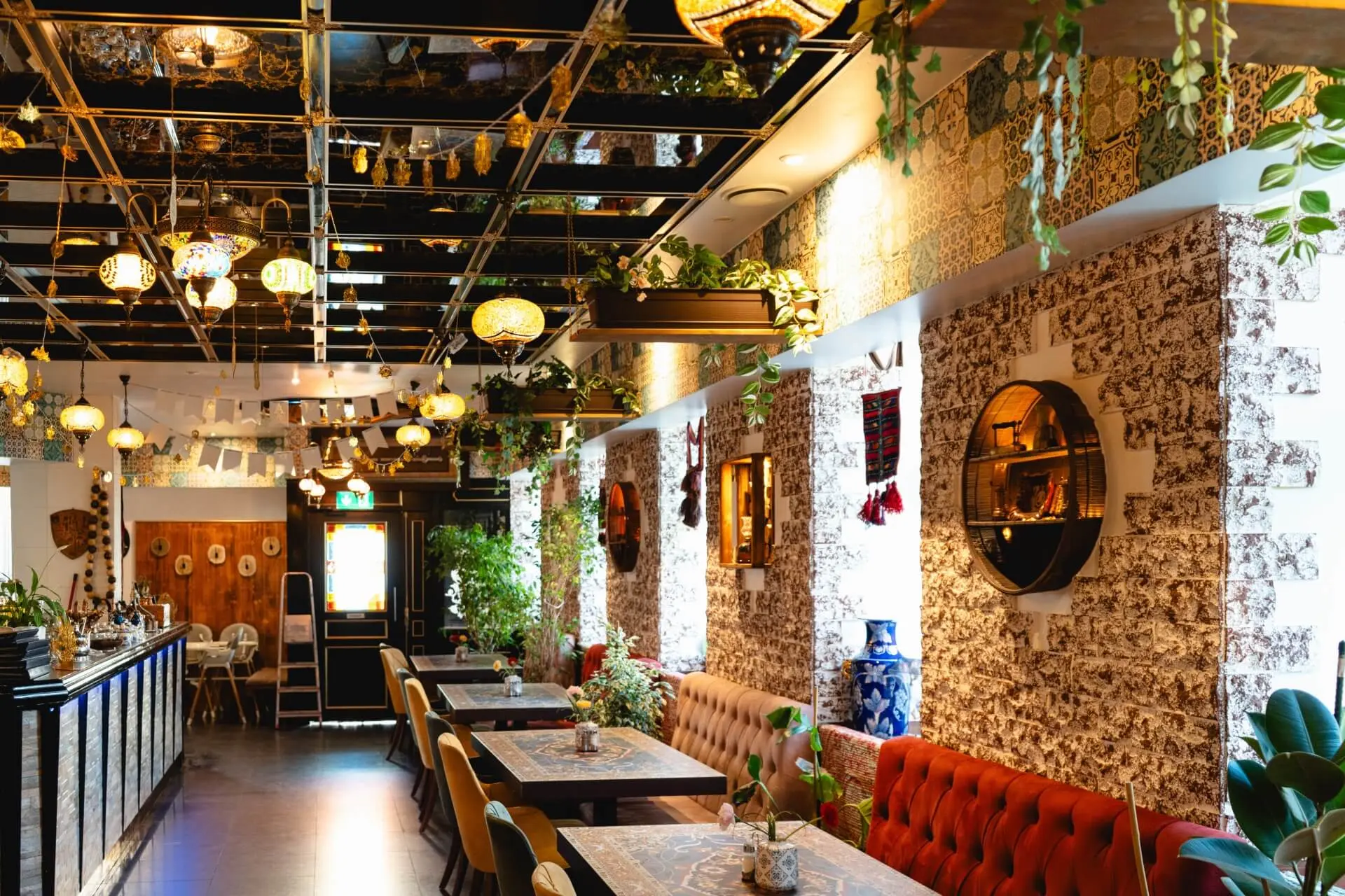 Ein lebhaftes Restaurantinterieur mit gemusterten Wänden, hängenden Pflanzen, runden Spiegeln, bunten Hängelampen und Tischen mit roten Sitzkissen, die eine gemütliche und einladende Atmosphäre schaffen.