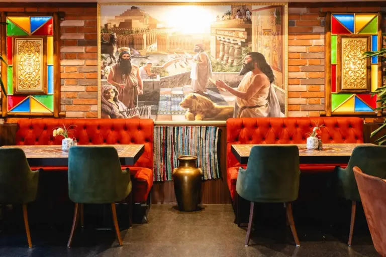 Ein gemütliches Restaurant mit Stühlen aus rotem und grünem Samt, Holztischen, bunten Glasfenstern und einem großen Wandgemälde mit antiken Figuren und einem Löwen an einer Backsteinwand. Unter dem Wandbild befindet sich eine dekorative Vase.