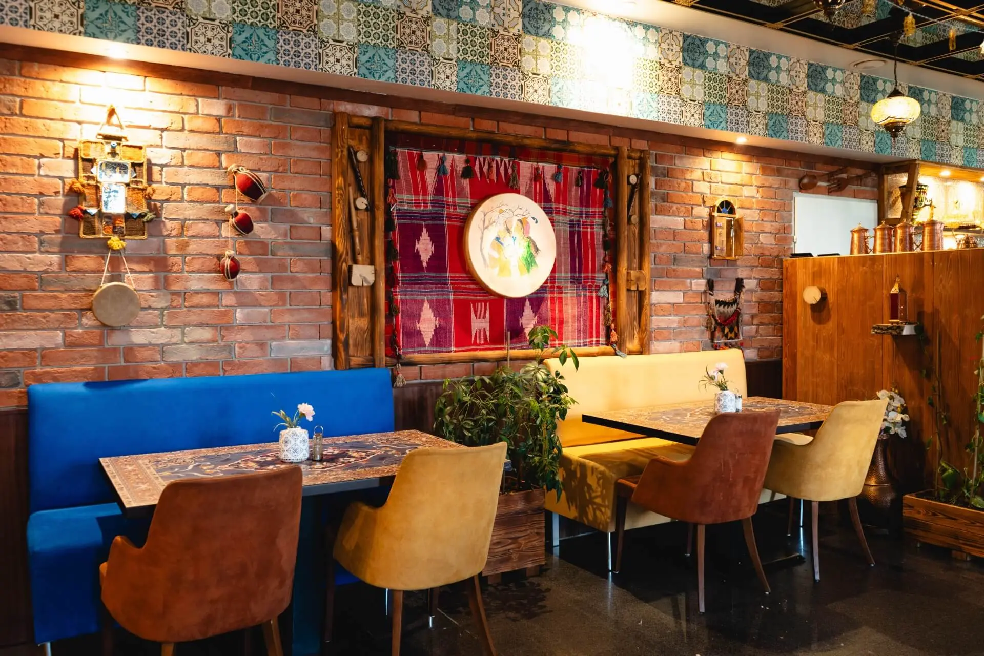 Ein gemütliches Restaurant mit Backsteinwänden, bunten Kacheln, blauen und gelben Sitzgelegenheiten, Holztischen und einer eklektischen Einrichtung mit Wandteppichen, gerahmter Kunst und hängenden Ornamenten. Pflanzen sorgen für einen Hauch von Grün.
