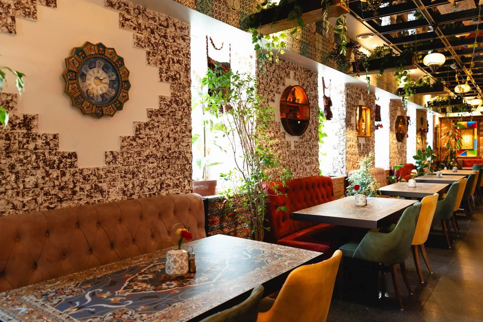 Ein gemütliches Restaurant mit gemusterten Wänden, bunt gepolsterten Stühlen und Sofas, Wandkunst, dekorativen Spiegeln und Grünpflanzen in der Nähe von großen Fenstern, die natürliches Licht hereinlassen.