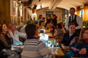 Eine lebhafte Restaurantszene mit Menschen, die an Tischen sitzen, sich unterhalten und ihr Essen genießen, während ein Kellner in der Nähe steht. Der Raum ist warm beleuchtet und bietet eine gemütliche, geschäftige Atmosphäre.