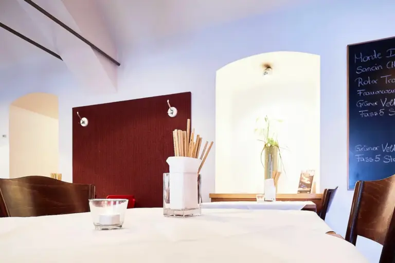 Ein weißer Tisch in einem hellen Restaurant mit einer Kerze, einem Glasbehälter mit Brotstangen und einer Pflanze im Hintergrund. Modernes Dekor mit roten und weißen Wänden und einer Kreidetafel als Speisekarte auf der rechten Seite.