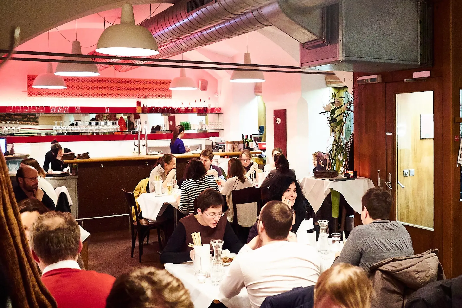 Ein belebtes Restaurant mit Menschen, die an Tischen sitzen, sich unterhalten und essen. Das Interieur zeichnet sich durch warme Beleuchtung, weiße Tischdecken und einen Barbereich im Hintergrund mit Gläsern und Flaschen in Regalen aus.