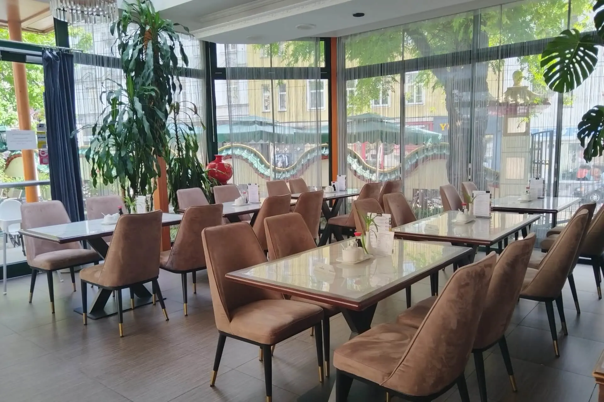 Ein helles, modernes Café mit Marmortischen, rosa gepolsterten Stühlen, Topfpflanzen und großen Fenstern mit Blick auf die Straße und die Bäume draußen. Der Raum wirkt sauber, einladend und menschenleer.