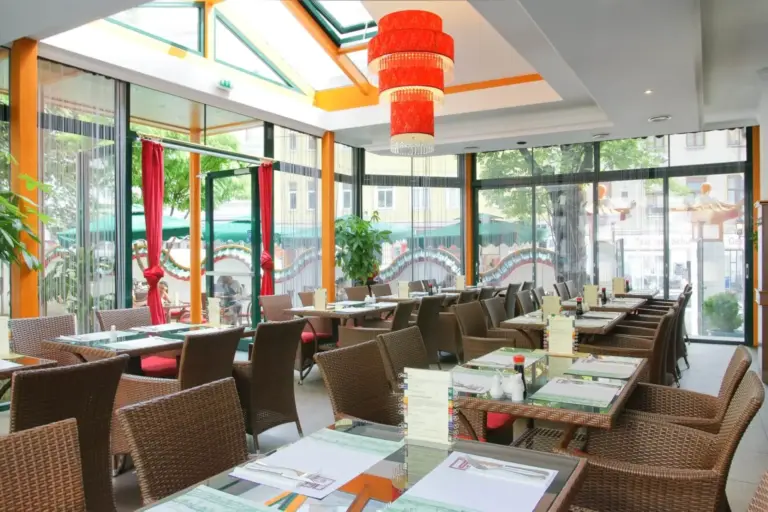 Ein helles, modernes Restaurant mit Glaswänden und -decken, roten Hängelampen, Korbsesseln und gedeckten Tischen. Durch die Fenster sind Grünflächen und Außenplätze zu sehen.