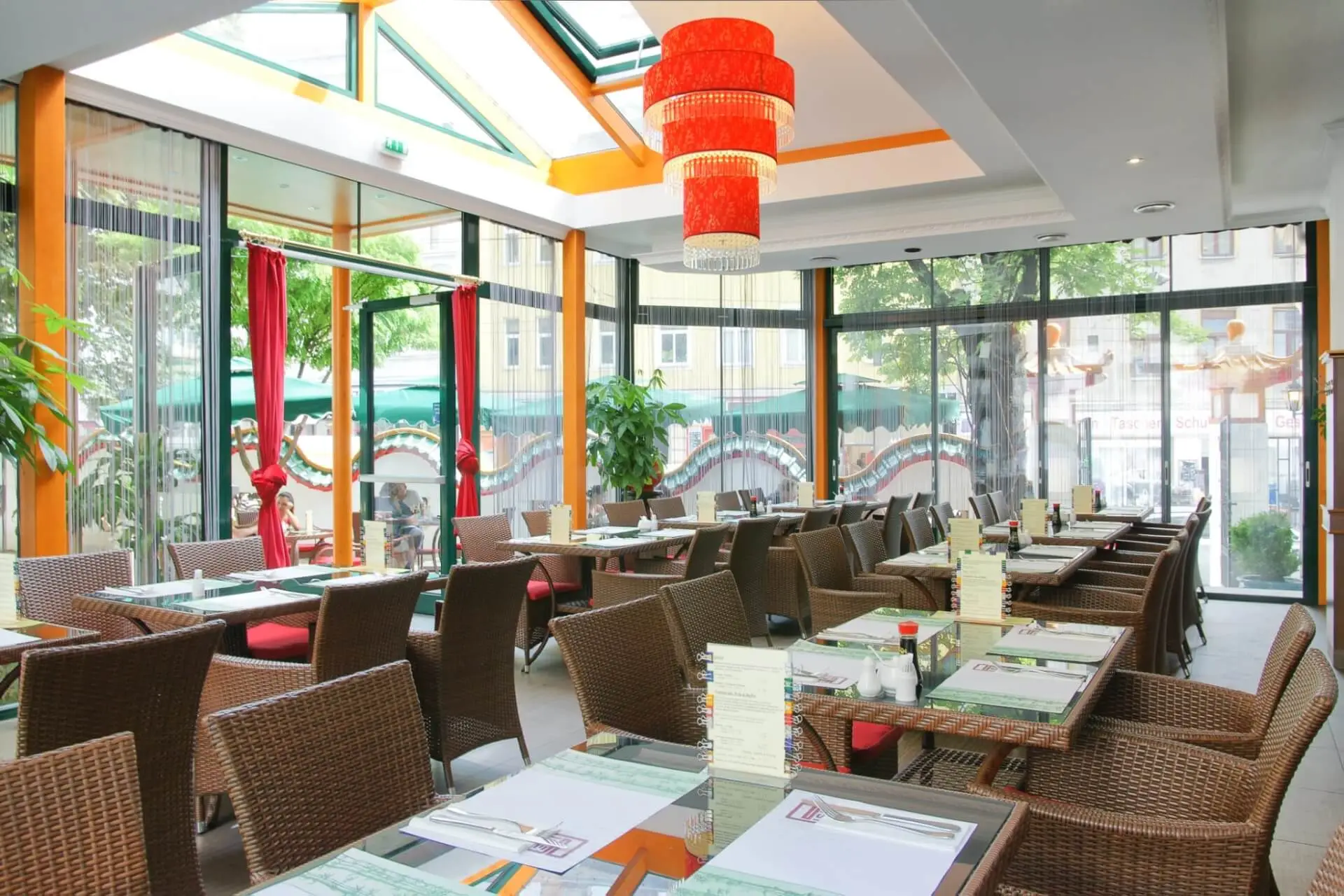 Ein helles, modernes Restaurant mit Glaswänden und -decken, roten Hängelampen, Korbsesseln und gedeckten Tischen. Durch die Fenster sind Grünflächen und Außenplätze zu sehen.