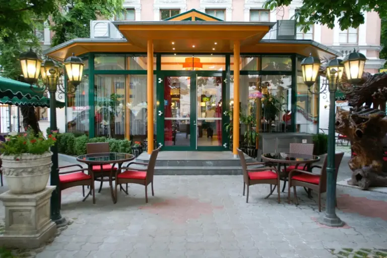 Sitzgelegenheiten im Freien mit rot gepolsterten Stühlen und runden Tischen auf einer Terrasse vor einem Restaurant mit großen Glasfenstern, gelben Säulen und dekorativen Lampen, umgeben von Grünflächen.