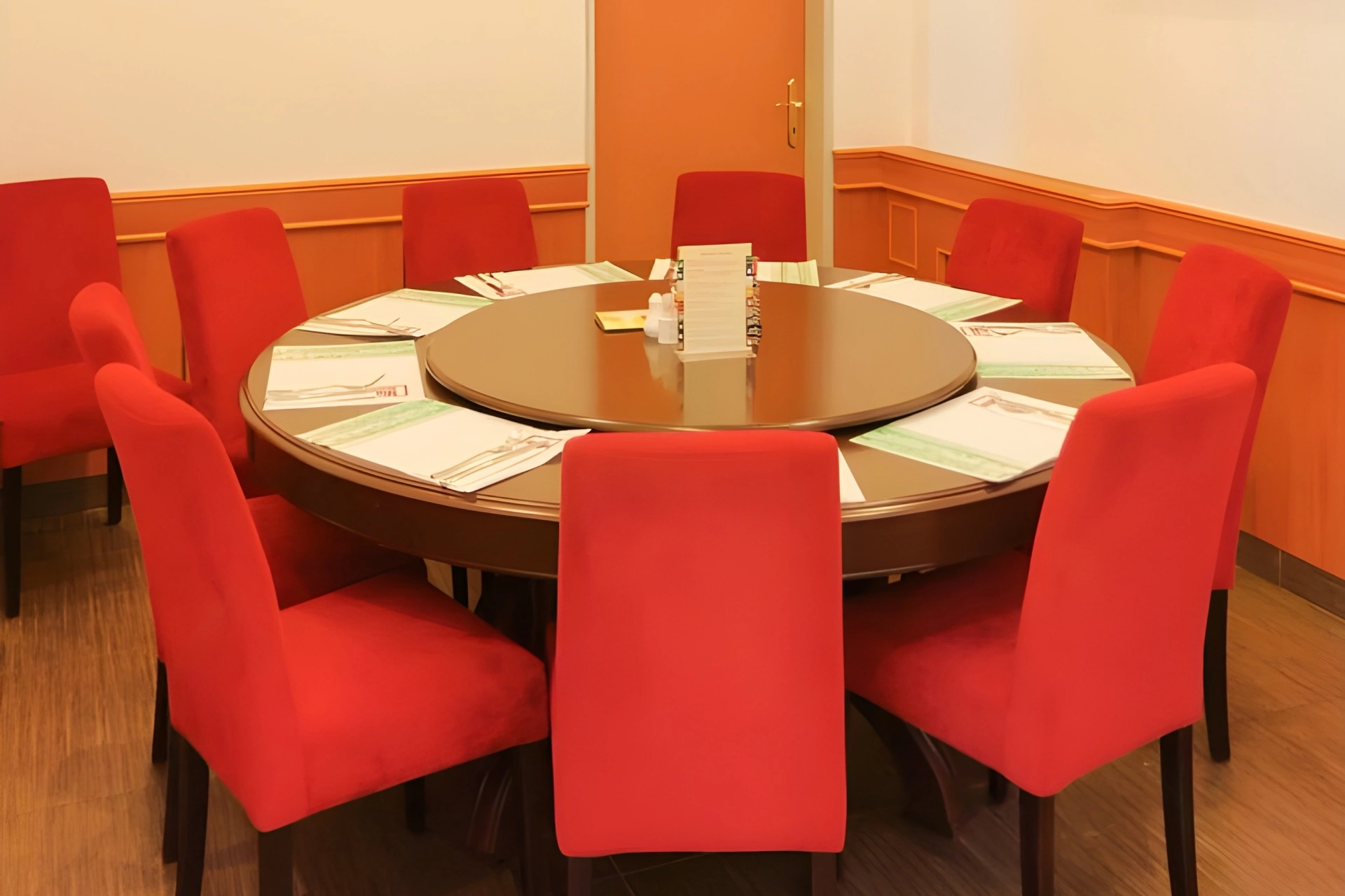 Ein runder Esstisch mit einer gläsernen Lazy Susan ist von acht roten Stühlen umgeben. Jeder Platz ist mit einem Tischset, einer Serviette und Besteck ausgestattet. Der Raum hat helle Wände und einen Holzfußboden.