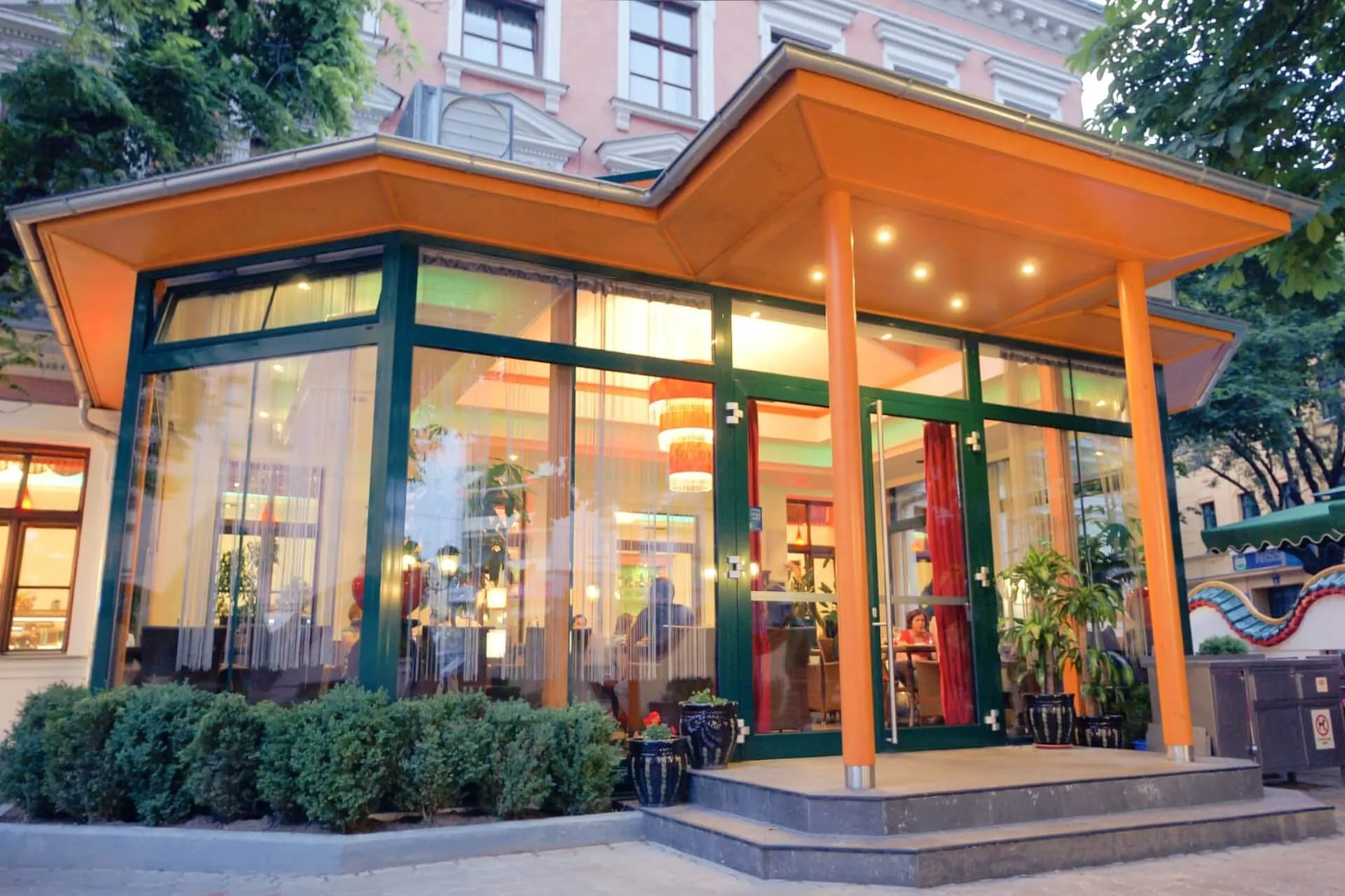 Ein modernes Restaurant mit großen Glasfenstern und einem orangefarbenen Dach, das innen warm beleuchtet ist. Der Eingang ist begrünt, und durch die Fenster kann man Menschen beim Essen beobachten. Das Gebäude befindet sich in einer städtischen Kulisse.