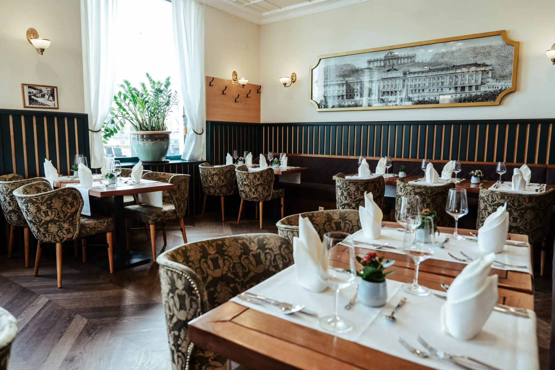 Eleganter Speisesaal des Restaurants mit Holztischen, gemusterten Polsterstühlen, weißen Servietten, Glaswaren, Topfpflanzen, großem Fenster mit Vorhängen, gerahmter Kunst an der Wand und geschmackvoller, klassischer Einrichtung.