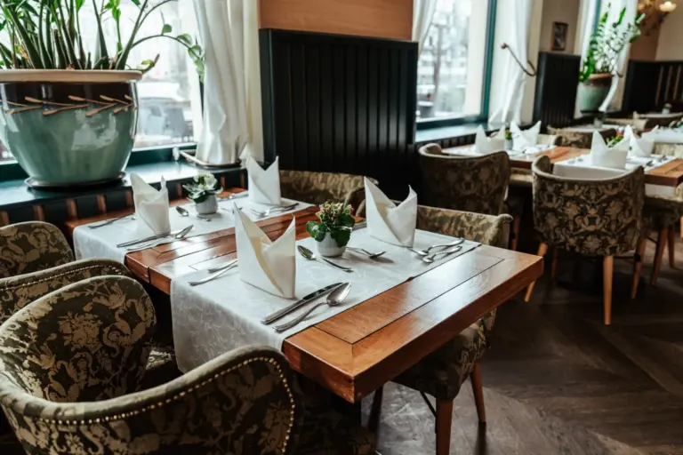 Ein fein säuberlich gedeckter Restauranttisch mit gefalteten weißen Servietten, Silberbesteck und kleinen Topfpflanzen, umgeben von verzierten Polsterstühlen, neben großen Fenstern mit weißen Vorhängen und grünen Zimmerpflanzen.