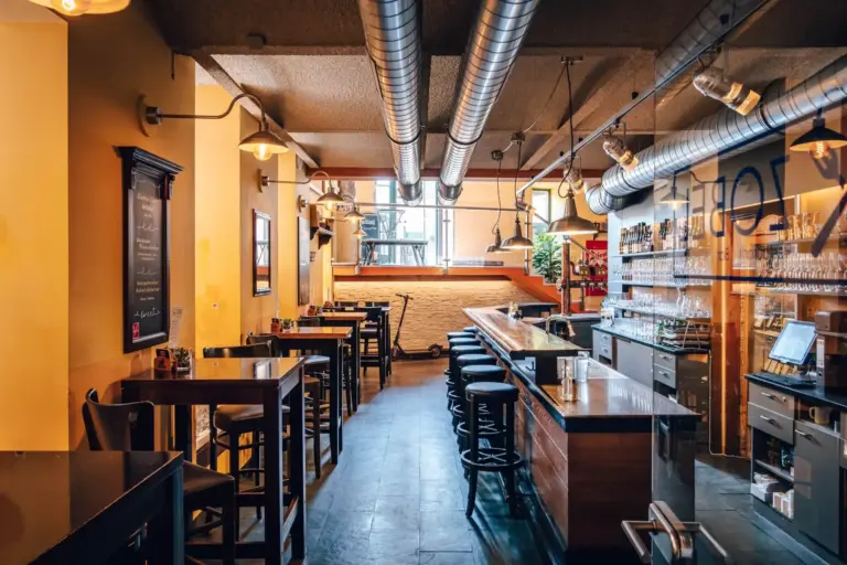 Ein modernes Café mit gelben Wänden, Hängelampen, Holztischen und -hockern, einem langen Bartresen, Regalen mit Flaschen und großen Luftschächten an der Decke. Durch ein Fenster an der Rückseite fällt Tageslicht ein.