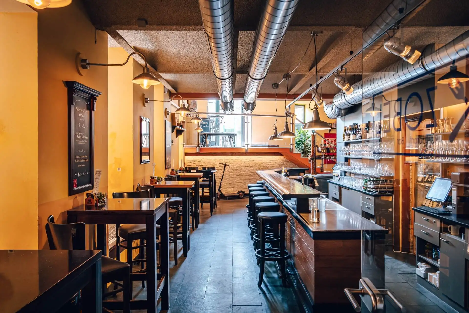 Ein modernes Café mit gelben Wänden, Hängelampen, Holztischen und -hockern, einem langen Bartresen, Regalen mit Flaschen und großen Luftschächten an der Decke. Durch ein Fenster an der Rückseite fällt Tageslicht ein.
