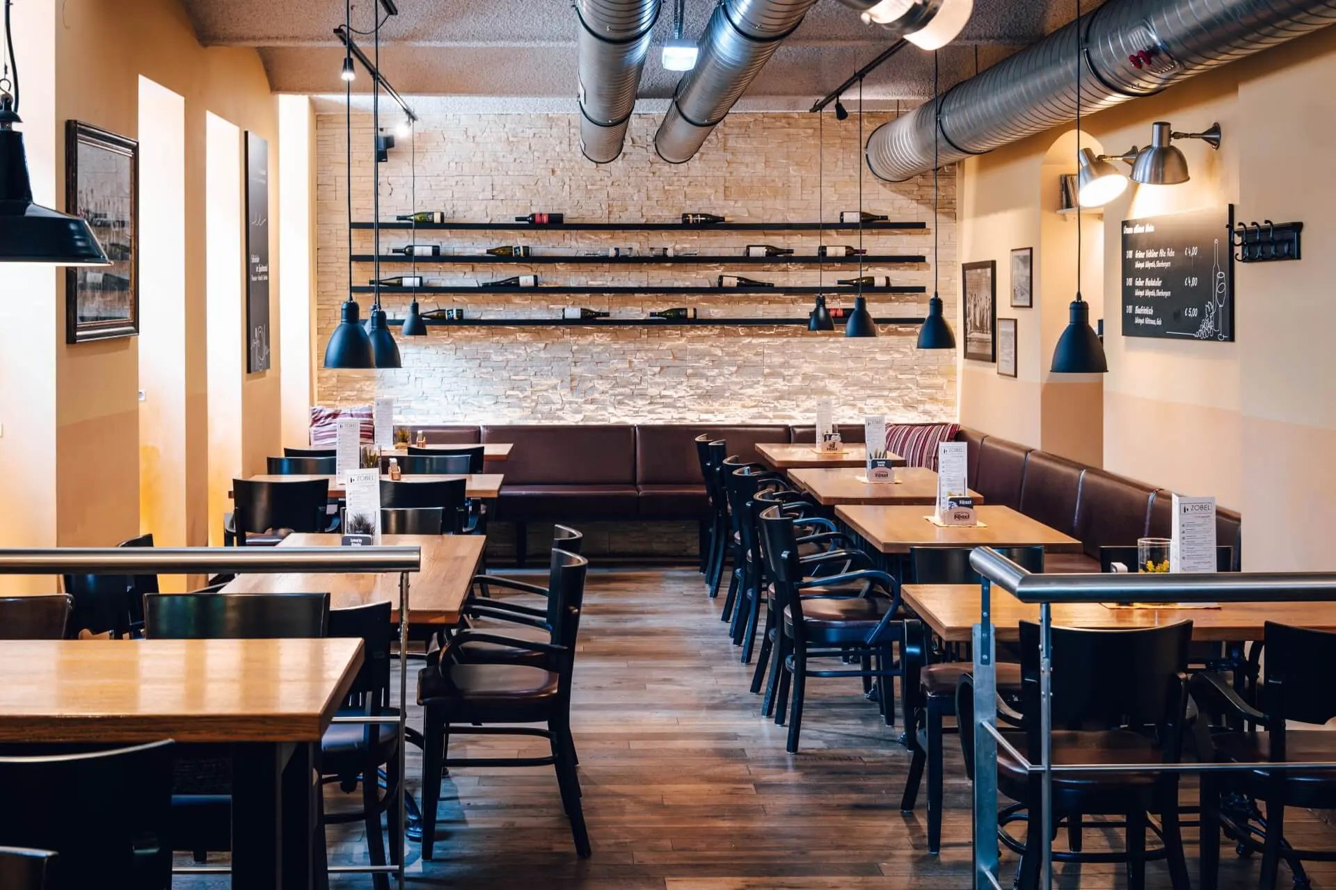 Ein modernes, gemütliches Restaurant mit Holztischen, schwarzen Stühlen, Beleuchtung im Industriestil und freiliegenden Luftschächten. An der Rückwand stehen Regale mit Dekoration, und auf jedem Tisch liegen Speisekarten. Der Raum ist menschenleer.