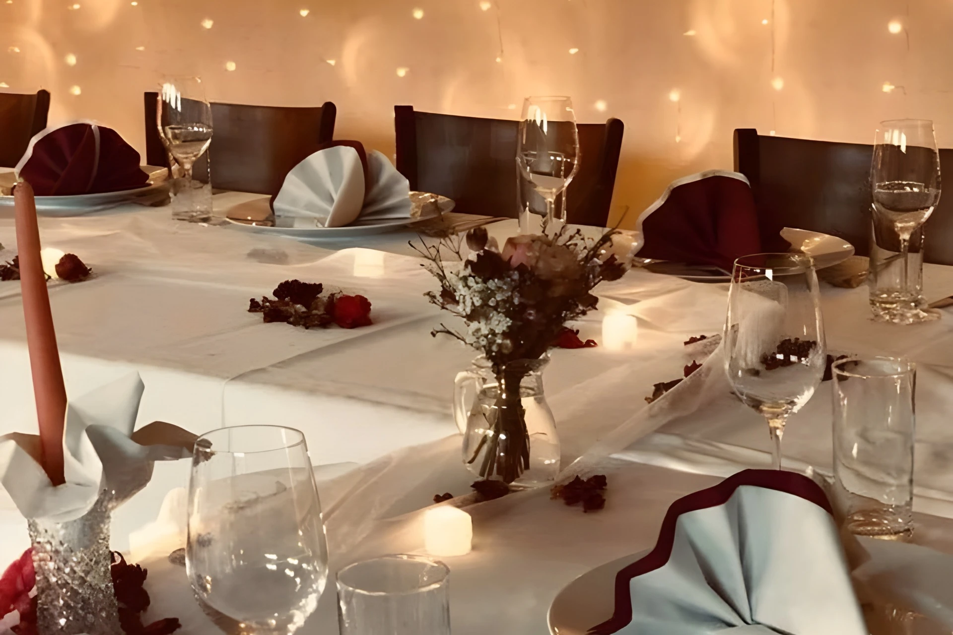 Elegant gedeckter Tisch mit gefalteten Servietten, Weingläsern und einem kleinen floralen Tafelaufsatz. Im Hintergrund hängen sanfte Lichterketten, und kleine Kerzen und Rosenblätter schmücken das weiße Tischtuch.