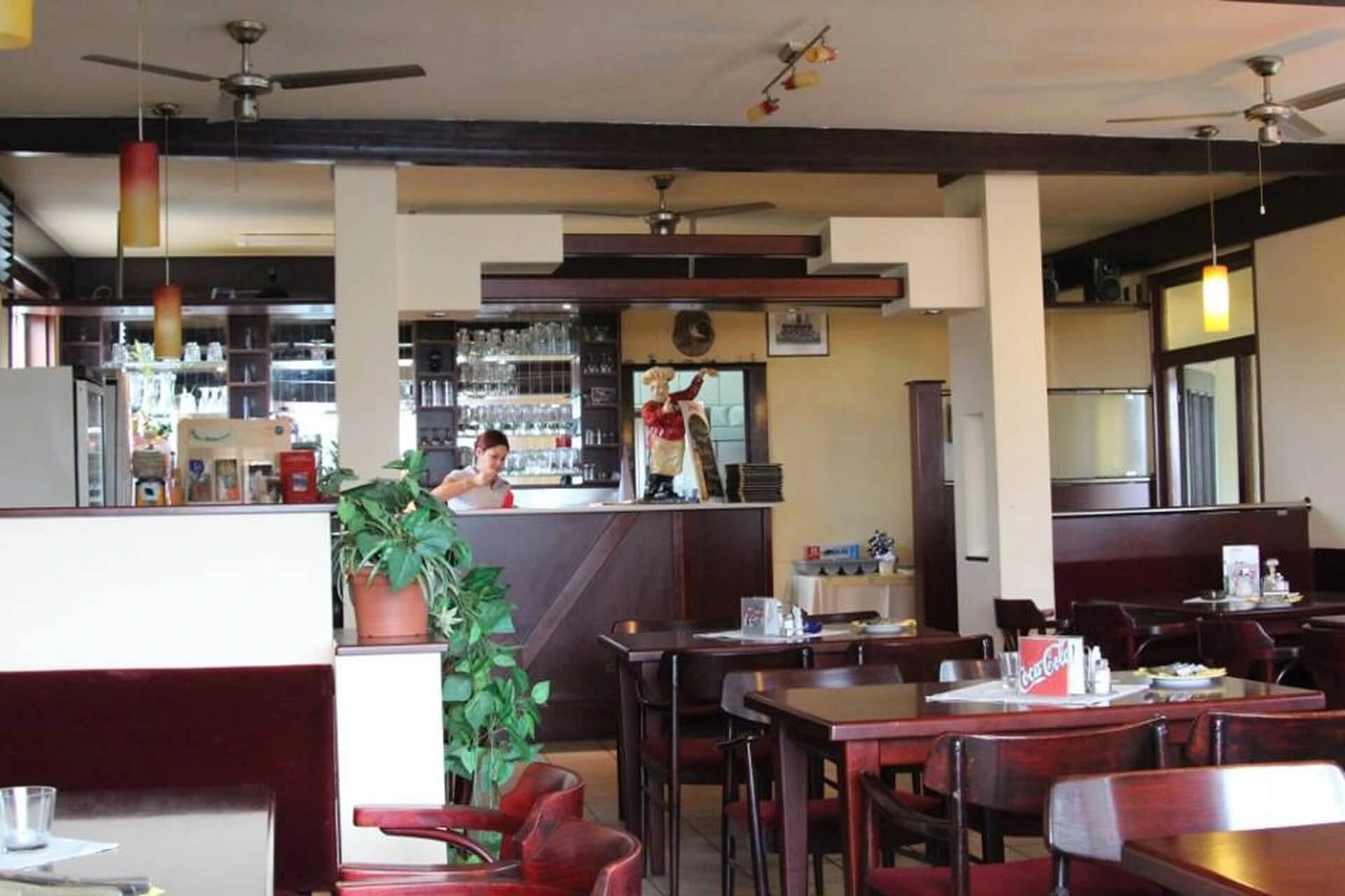 Ein gemütliches Restaurant mit Holztischen und -stühlen, einer Topfpflanze und einem Barbereich im Hintergrund, in dem eine Person Getränke zubereitet. Der Raum wirkt sauber und gut beleuchtet mit natürlichem und künstlichem Licht.