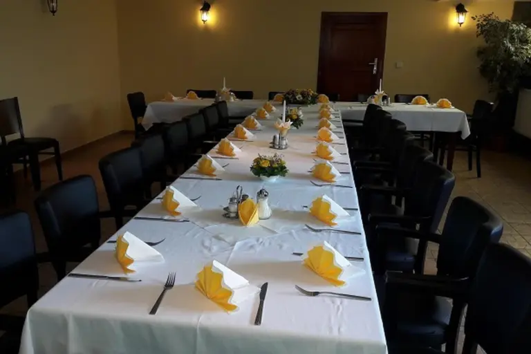 Ein langer Esstisch mit weißem Tischtuch, gedeckt mit gelben und weißen gefalteten Servietten, Besteck und kleinen Blumensträußen, umgeben von schwarzen Stühlen in einem gut beleuchteten Raum.