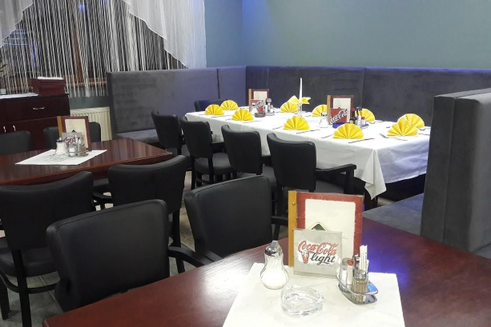 Ein Restaurant mit gedeckten Tischen, grauen gepolsterten Stühlen, schwarzen Stühlen und weißen Tischtüchern. Gelbe Servietten sind ordentlich gefaltet, und auf den Tischen liegen Coca-Cola Light-Menüs aus.