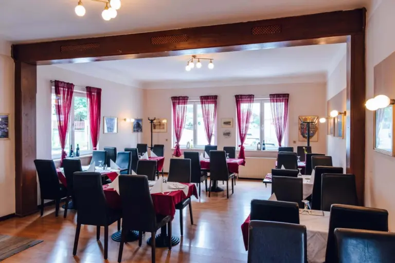 Ein heller, moderner Speisesaal mit schwarzen Stühlen und Tischen, die mit weißen und roten Tischdecken bedeckt sind. Große Fenster mit roten Vorhängen lassen natürliches Licht herein, und Wandkunst schmückt den Raum.