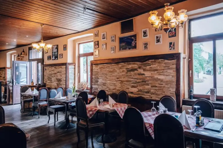 Gemütliches Restaurantinterieur mit Holzdecke, Vintage-Kronleuchtern und gerahmten Fotos an den Wänden. Die Tische sind mit rotkarierten Tischdecken bedeckt und mit Gläsern und Servietten gedeckt. Große Fenster lassen viel natürliches Licht herein.