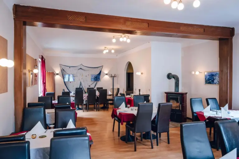 Ein Restaurant mit schwarzen Stühlen, rot-weiß gedeckten Tischen, Holzböden, einem Holzofen und Wandleuchten. An der Rückwand hängt ein dekoratives Fischernetz.