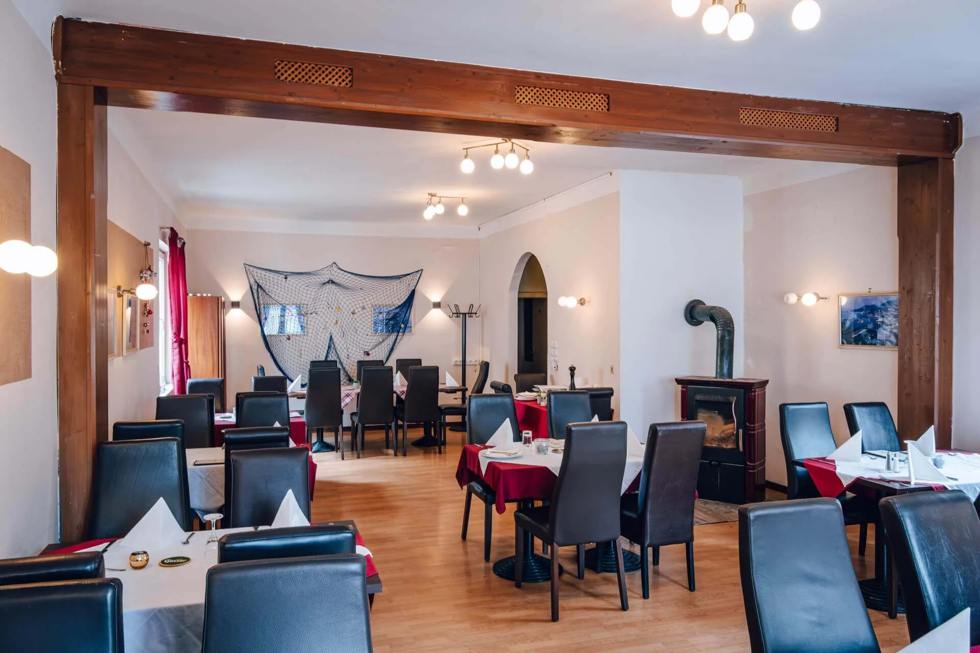 Ein Restaurant mit schwarzen Stühlen, rot-weiß gedeckten Tischen, Holzböden, einem Holzofen und Wandleuchten. An der Rückwand hängt ein dekoratives Fischernetz.