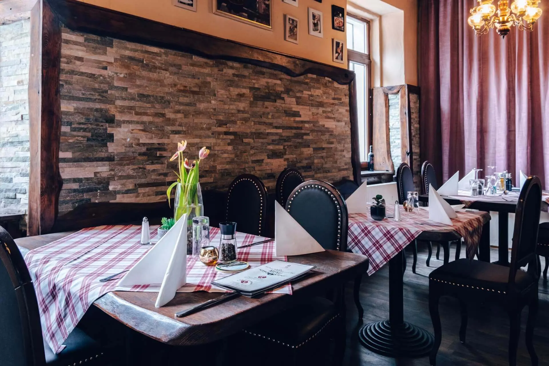 Ein gemütliches Restaurant mit karierten Tischtüchern, schwarzen Stühlen und ordentlich gefalteten Servietten auf den Tischen. Es gibt Blumen in Vasen, Akzentwände aus Stein, gerahmte Bilder und große Fenster, die natürliches Licht hereinlassen.