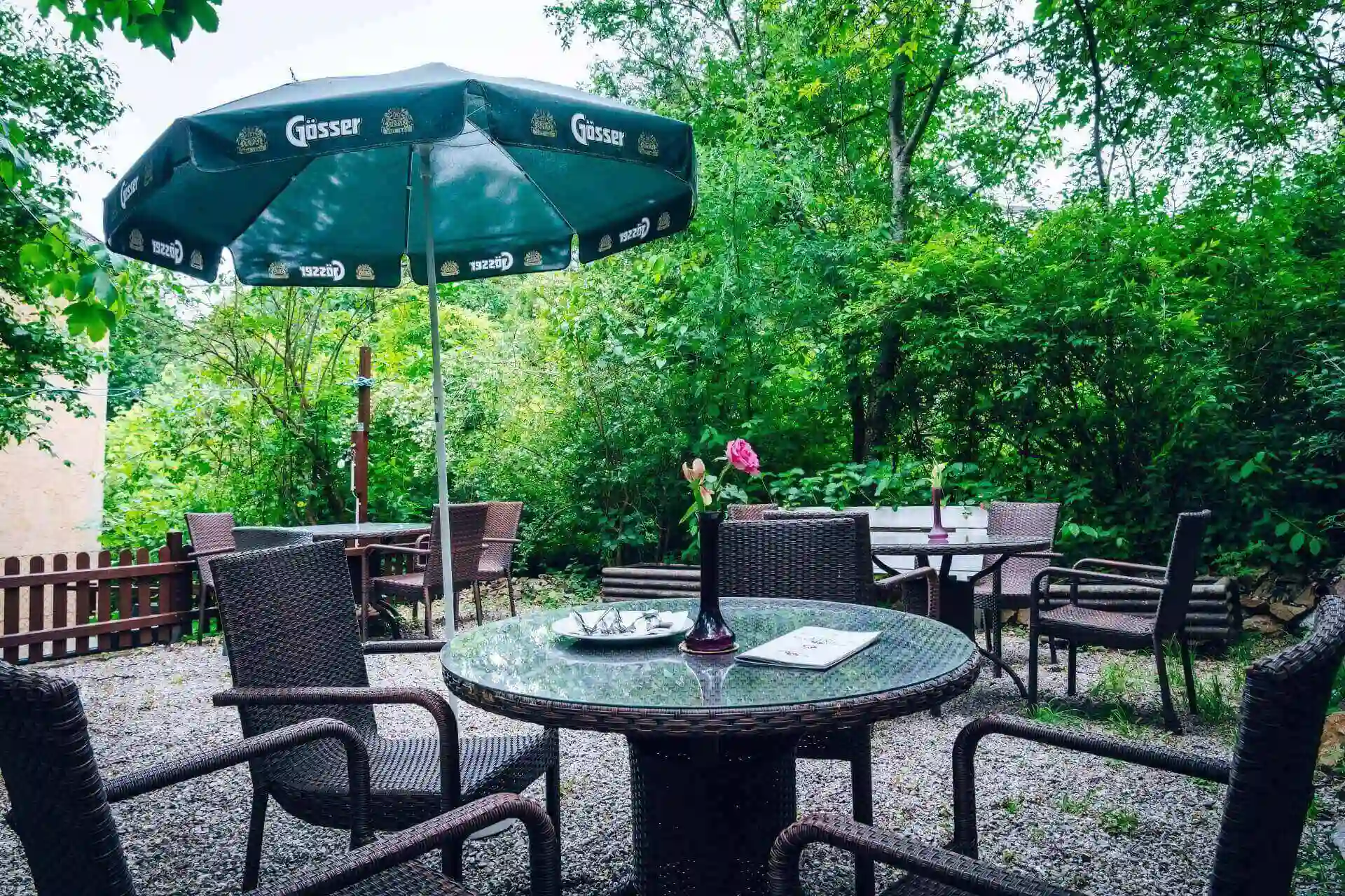 Terrasse im Freien mit Korbstühlen um einen Glastisch, einem Blumenvasen-Mittelstück und einem großen grünen Sonnenschirm. Umgeben von üppigen grünen Bäumen und Sträuchern, die eine gemütliche, einladende Gartenatmosphäre schaffen.
